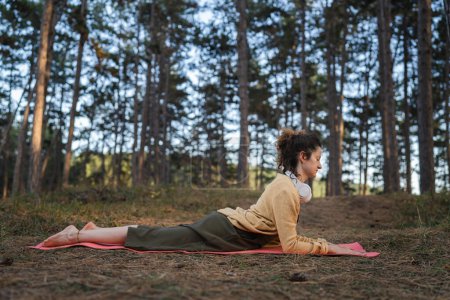 Téléchargez les photos : Une jeune femme adulte caucasienne couchée seule dans le parc ou dans la forêt dans la nature avec écouteurs pour la méditation guidée en ligne équilibre mental et pratiques de manifestation concept - en image libre de droit