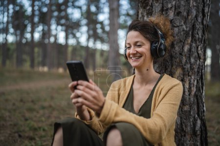 Foto de Una mujer joven adulta caucásica se sienta sola en el parque o bosque en la naturaleza con auriculares preparándose para la meditación guiada auto-cuidado manifestación práctica mental equilibrio emocional concepto - Imagen libre de derechos