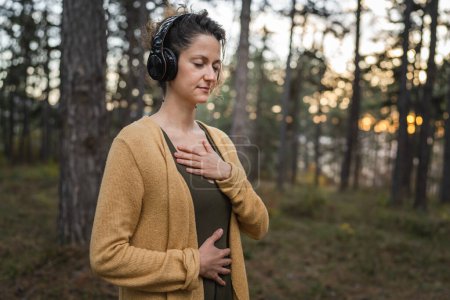 Photo for One woman young adult caucasian female stand alone in the park or forest in nature with headphones preparing for guided meditation self-care manifestation practice mental emotional balance concept - Royalty Free Image