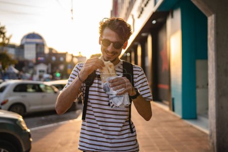Téléchargez les photos : Un homme jeune adulte caucasien moderne mâle dans la ville en journée ensoleillée marcher et manger sandwich fast food concept vie urbaine copier l'espace touristique manger dans la rue personne réelle - en image libre de droit