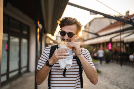 Téléchargez les photos : Un homme jeune adulte caucasien moderne mâle dans la ville en journée ensoleillée marcher et manger sandwich fast food concept vie urbaine copier l'espace touristique manger dans la rue personne réelle - en image libre de droit