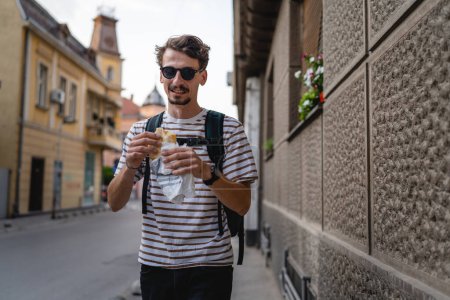 Photo for One man young adult modern caucasian male in the city in sunny day walk and eat sandwich fast food concept urban life copy space tourist eating on the street real person - Royalty Free Image