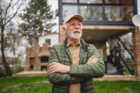 Photo for One modern senior caucasian man portrait of male grandfather with beard and hat cap stand in front of his house in day wear jacket happy confident copy space arms crossed - Royalty Free Image
