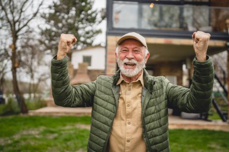 Téléchargez les photos : Un homme caucasien senior moderne portrait de grand-père masculin avec barbe et chapeau se tenir devant sa maison en veste de jour heureux espace de copie confiant célébrer victoire - en image libre de droit
