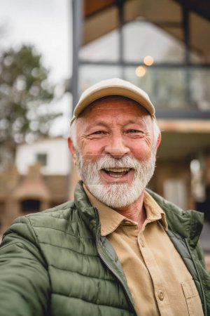 Téléchargez les photos : Un homme caucasien supérieur moderne portrait de grand-père masculin avec barbe et chapeau cap stand en face de sa maison dans la veste de jour heureux espace de copie confiant - en image libre de droit
