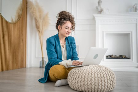 Photo for One woman young adult caucasian female sitting on the floor at home with laptop computer working and looking to the side happy smile freelance brunette with curly hair domestic life copy space - Royalty Free Image