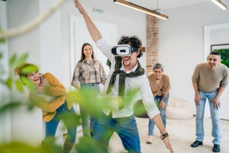 Téléchargez les photos : Un homme adulte caucasien mâle devant un groupe d'hommes et de femmes amis profiter de réalité virtuelle casque VR au travail en s'amusant ensemble pendant le séminaire de team building vraies personnes filtre lumineux - en image libre de droit