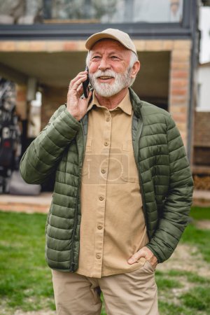 Téléchargez les photos : Un homme homme caucasien senior avec barbe grise et casquette debout en plein air au printemps ou en automne jour heureux sourire utiliser téléphone intelligent mobile pour faire un appel parler heureux sourire copier l'espace vraies personnes - en image libre de droit