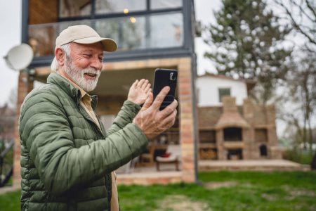 Téléchargez les photos : Un homme homme caucasien senior avec barbe grise et casquette debout en plein air au printemps ou en automne jour heureux sourire utiliser téléphone intelligent mobile pour la maison d'appel vidéo espace de copie personnes réelles - en image libre de droit