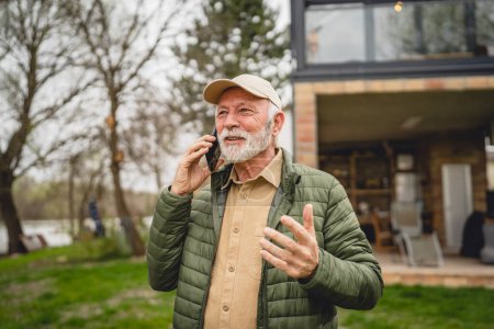 Photo for One man senior caucasian male with gray beard and cap stand outdoor in spring or autumn day happy smile use mobile smart phone to make a call talk happy smile excited copy space real people - Royalty Free Image