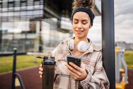 Photo for One woman young adult generation z caucasian modern female with headphones and supplement shaker happy smile use mobile phone app for training or music browse online in the city real person copy space - Royalty Free Image