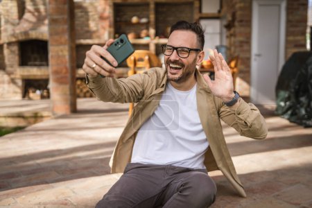 Foto de Un hombre feliz caucásico uso masculino smartphone para selfie foto o videollamada al aire libre en frente de la pequeña casa moderna, mientras que en vacaciones en el día de la gente real copiar espacio - Imagen libre de derechos