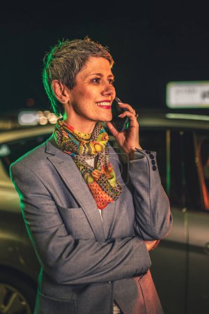 Téléchargez les photos : Une femme âgée cheveux gris court caucasien femme utiliser téléphone mobile pour parler ou passer un appel dans la ville la nuit debout devant la voiture heureux sourire confiant espace de copie personne réelle - en image libre de droit