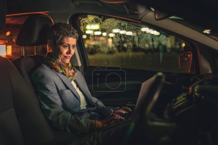 Photo for One mature senior woman caucasian female sitting in the car working on laptop computer at night in dark with short gray hair modern manager or business owner on road copy space - Royalty Free Image