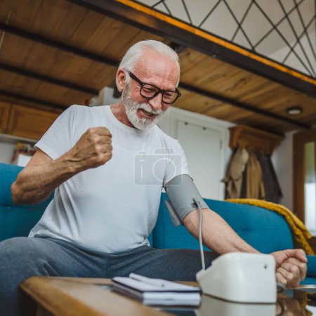Téléchargez les photos : Un homme heureux homme caucasien âgé avec la barbe en bonne santé célébrer utiliser appareil de pression artérielle pour vérifier et mesurer les résultats tout en s'asseyant à la table à la maison seul les gens réels copier espace concept de soins de santé - en image libre de droit
