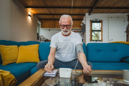 Téléchargez les photos : Un homme homme caucasien âgé avec barbe utiliser un appareil de pression artérielle pour vérifier et mesurer les résultats tout en s'asseyant à la table à la maison seul les gens réels copier l'espace concept de soins de santé - en image libre de droit