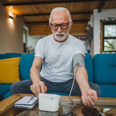 Photo for One man senior caucasian male with beard use blood pressure device to check and measure results while sit at the table at home alone real people copy space health care concept - Royalty Free Image