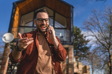 Téléchargez les photos : Un homme adulte caucasien mâle avec barbe et lunettes de vue debout en plein air dans la journée ensoleillée sourire heureux utiliser téléphone intelligent mobile pour faire un appel parler copie espace vraies personnes - en image libre de droit