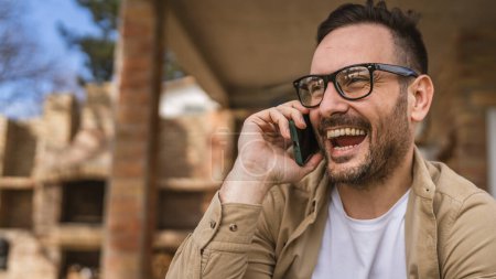 Téléchargez les photos : Un homme adulte caucasien mâle avec barbe et lunettes de vue assis en plein air dans la journée ensoleillée sourire heureux utiliser téléphone intelligent mobile pour faire un appel parler copie espace vraies personnes - en image libre de droit