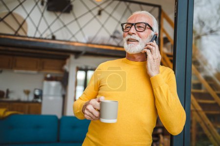 Téléchargez les photos : Un homme homme caucasien senior avec barbe grise debout sur le balcon à la porte dans la journée sourire heureux utiliser téléphone intelligent mobile pour faire un appel parler copie espace tenir tasse de café matin routine quotidienne - en image libre de droit