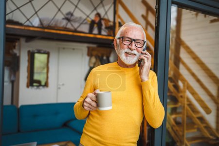 Foto de Un hombre mayor de sexo masculino caucásico con la barba gris de pie en el balcón en la puerta en el día sonrisa feliz uso de teléfono inteligente móvil para hacer una llamada hablar espacio copia taza de café por la mañana rutina diaria - Imagen libre de derechos