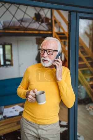 Foto de Un hombre mayor caucásico macho con la barba gris de pie en el balcón en la puerta en el día el uso de teléfono inteligente móvil para hacer una llamada hablar espacio de copia mantenga taza de café preocupado reflexivo - Imagen libre de derechos