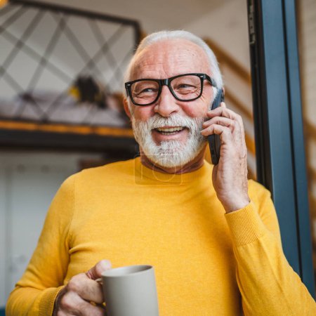 Foto de Un hombre mayor de sexo masculino caucásico con la barba gris de pie en el balcón en la puerta en el día sonrisa feliz uso de teléfono inteligente móvil para hacer una llamada hablar espacio copia taza de café por la mañana rutina diaria - Imagen libre de derechos