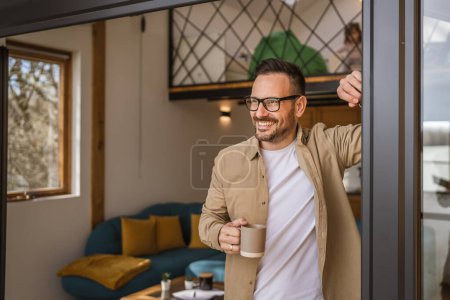 Téléchargez les photos : Un homme adulte caucasien mâle avec barbe et lunettes debout sur le balcon à la porte dans la journée heureux sourire tenir tasse de café copie espace matin routine quotidienne concept - en image libre de droit