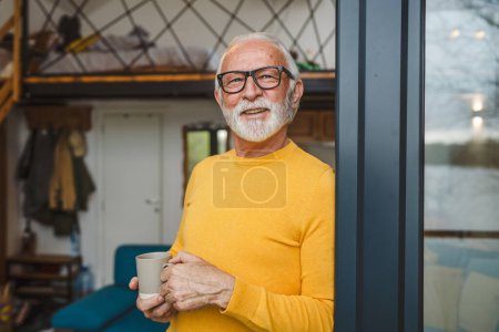 Foto de Un hombre mayor caucásico macho con barba y gafas de pie en el balcón en la puerta en el día sonrisa feliz sostener taza de café copia espacio mañana concepto de rutina diaria - Imagen libre de derechos