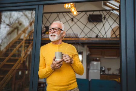 Téléchargez les photos : Un homme homme caucasien senior avec barbe et lunettes debout sur le balcon à la porte dans la journée heureux sourire tenir tasse de café copie espace matin routine quotidienne concept - en image libre de droit