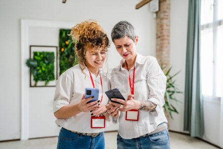 Photo for Two women caucasian mature female colleagues use smartphone while take a brake from work browse internet or social network app talk chat relax happy smile real people copy space bright photo - Royalty Free Image