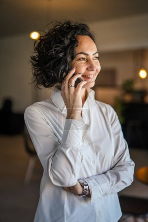 Photo for One woman mature caucasian female businesswoman entrepreneur stand at work or home use mobile phone making a call talk real people copy space wear white shirt curly hair happy smile - Royalty Free Image