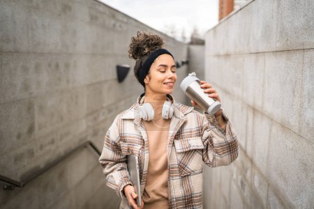 Foto de Una mujer joven adulta estudiante de pie o caminar al aire libre sostener ordenador portátil y termo taza de café persona real copiar espacio sonrisa feliz confiado con auriculares en el día - Imagen libre de derechos