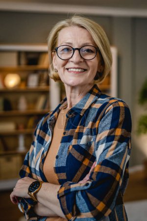 Téléchargez les photos : Portrait d'une femme caucasienne blonde mature avec des lunettes à la maison sourire heureux regardant vers l'appareil photo confiant porter l'espace de copie de chemise - en image libre de droit