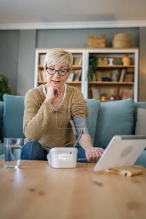 Photo for One woman senior caucasian female use blood pressure device on hand to check health results while sit at home real people health care concept - Royalty Free Image