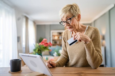 Téléchargez les photos : Une femme âgée mature grand-mère s'asseoir à la maison utiliser carte de crédit ou de débit pour les achats en ligne parcourir les magasins en ligne acheter des trucs utiliser tablette numérique personnes réelles copier l'espace - en image libre de droit