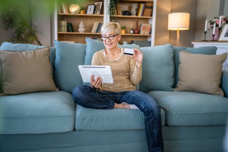 Téléchargez les photos : Une femme âgée mature grand-mère s'asseoir à la maison utiliser carte de crédit ou de débit pour les achats en ligne parcourir les magasins en ligne acheter des trucs utiliser tablette numérique personnes réelles copier l'espace - en image libre de droit