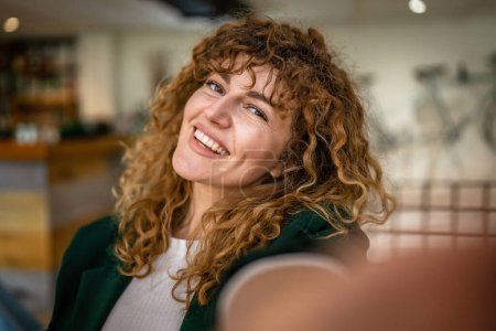 Foto de Una mujer caucásica mujer feliz seguro de pie interior en la cafetería con el pelo rizado sonrisa persona real copia espacio autorretrato selfie UGC usuario contenido generado - Imagen libre de derechos