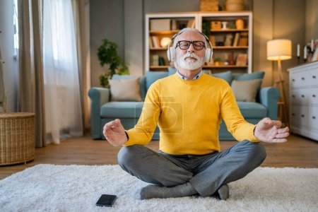 Téléchargez les photos : Un homme mature homme caucasien aîné utilisant des écouteurs pour la méditation guidée en ligne pratiquant la pleine conscience manifestation du yoga avec les yeux fermés à la maison personnes réelles self care concept copier l'espace - en image libre de droit