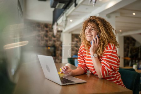 Téléchargez les photos : Une femme adulte femme caucasienne assis au travail de café sur l'ordinateur portable faire un appel téléphonique parler sourire heureux confiant souriant espace de copie personne réelle - en image libre de droit