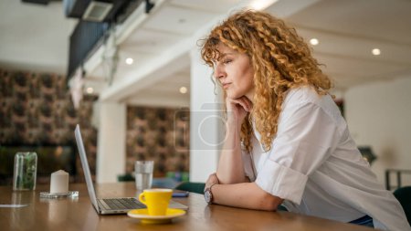 Téléchargez les photos : Une femme adulte caucasienne avec des cheveux bouclés travailler sur ordinateur portable tout en s'asseyant au café espace de copie personne réelle heureux sourire confiant - en image libre de droit