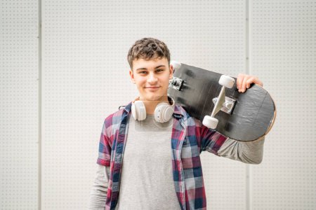 Photo for One man young adult caucasian teenager stand outdoor with skateboard on his shoulder and headphones posing portrait looking to the camera happy confident wear shirt casual real person copy space - Royalty Free Image