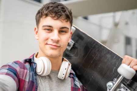 Photo for One man young adult caucasian teenager stand outdoor with skateboard on his shoulder and headphones posing portrait looking to the camera happy confident wear shirt casual real person copy space - Royalty Free Image
