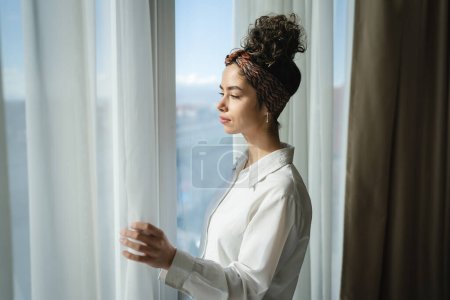 Photo for One woman beautiful caucasian female standing by the window at home or hotel room looking outside in sunny day wear white shirt copy space waiting alone - Royalty Free Image