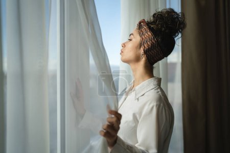 Photo for One woman beautiful caucasian female standing by the window at home or hotel room looking outside in sunny day wear white shirt copy space waiting alone - Royalty Free Image