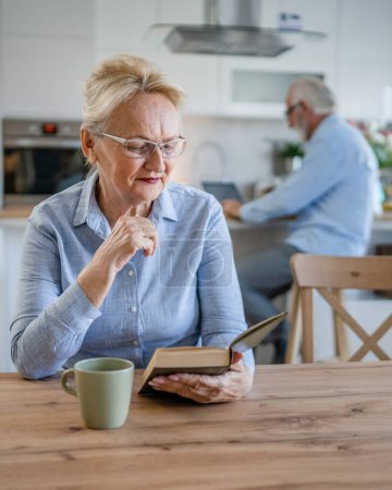 Photo for One woman mature senior caucasian female pensioner modern grandmother read book at home wear eyeglasses while her husband stand behind family life active senior real people copy space - Royalty Free Image