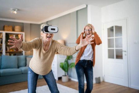 Foto de Dos mujeres maduras mayores caucásicas amigas o hermanas en casa disfrutar de realidad virtual VR auriculares personas reales activos senior divertirse concepto de ocio - Imagen libre de derechos