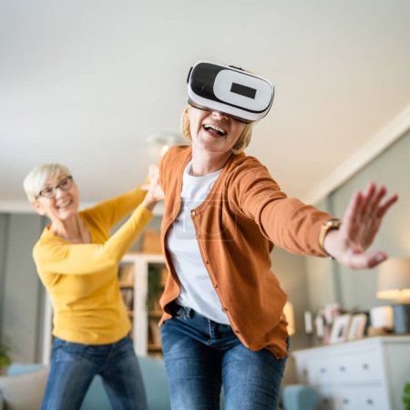 two women mature senior caucasian female friends or sisters at home enjoy virtual reality VR headset real people active senior having fun leisure concept