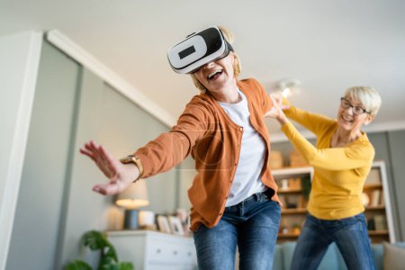 two women mature senior caucasian female friends or sisters at home enjoy virtual reality VR headset real people active senior having fun leisure concept