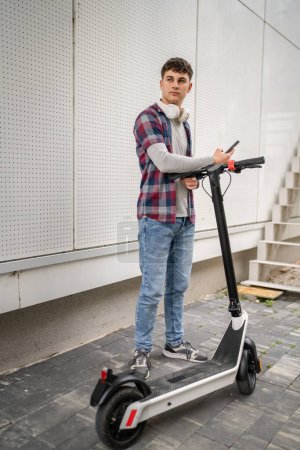 Photo for One young caucasian man male teenager generation Z with electric kick push scooter at modern building in the city eco mode of transport concept use mobile smart phone for sms text messages copy space - Royalty Free Image